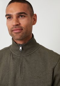 Suéter verde oliva con un cuello alto y media cremallera. Hecho de un tejido suave, con detalles de costura sutiles y un logo en el pecho.