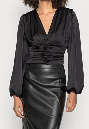 Femme portant un chemisier noir à manches longues plissé avec un profond décolleté en V, associé à une jupe en cuir noire taille haute, sur fond uni.