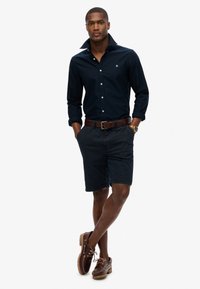 Marineblauwe overhemshirt met een verhoogde kraag, donkere shorts en bruine boot schoenen met veters. Inclusief een leren bruine riem en een polshorloge.