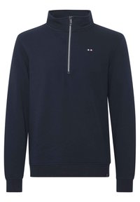 Pulover u tamnoplavoj boji s kvartnim patentnim zatvaračem, rebrastim manžetama i malim logotipom u crvenoj, bijeloj i plavoj boji na prsima. Glatka tekstura.