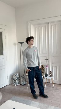 Fiatal férfi, göndör hajjal, szürke pulóvert és sötét nadrágot visel, egy világos szobában áll, fehér ajtókkal és dekoratív tárgyakkal.