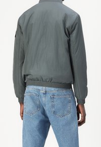 Graue Bomberjacke mit glatter Oberfläche, gerippten Bündchen und Saum, getragen über hellblauen Jeans mit klassischem Gesäßtaschendesign.