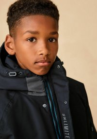 Schwarze wasserdichte Jacke mit hohem Kragen, verstellbarem Kapuze und blauen Reißverschlussakzenten. Das Material wirkt glatt mit glänzenden Beschlägen.