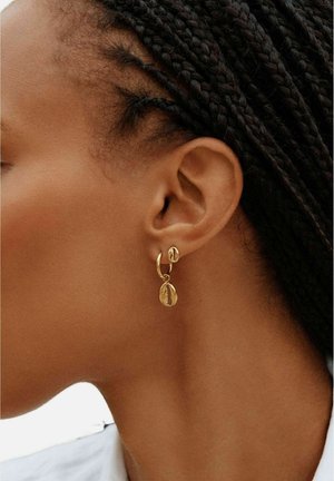 Seitenansicht einer Person mit geflochtenem Haar, die goldene Ohrringe in Form von Kaurimuscheln am linken Ohr trägt.