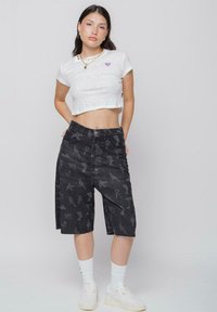 Camiseta blanca corta con gráfico de corazón morado; pantalones cortos de mezclilla negros de tiro alto con un patrón gris; modelo lleva zapatillas blancas y calcetines.