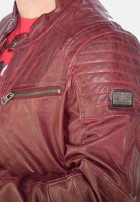 Rode leren jas met een glanzende afwerking, voorzien van doorgestikte schouderdetails, een ritssluiting zak en een getextureerde patch op de mouw.
