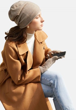 Brauner Wollmantel mit breiter Kragen, cremefarbener Rollkragenpullover, graue Handschuhe und hellblaue Jeans. Graue Mütze mit weicher Textur. Hält ein Smartphone.