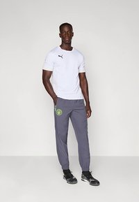 Bela majica s kratkimi rokavi in črnim logotipom, sive hlače z zelenim emblemom ter črni športni čevlji z otesano zasnovo in elastičnimi zapestji.