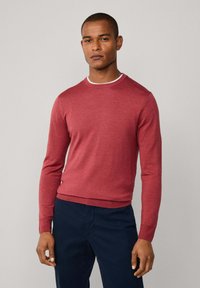 Muž v přiléhavém dlouhém červeném svetru přes bílou košili, v kombinaci s tmavě modrými kalhotami, stojící na jednolitém pozadí.