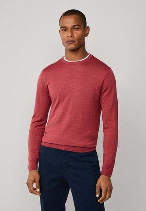 Uomo con indosso un maglione rosso attillato a maniche lunghe sopra una camicia bianca, abbinato a pantaloni blu scuro, in piedi contro uno sfondo neutro.