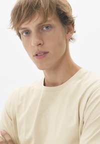 T-shirt beige clair à col rond, fabriqué en tissu doux, présentant une texture lisse et un éclat subtil. Le modèle a les cheveux courts et ébouriffés.