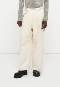 Pantaloni din denim bej, supradimensionați, cu croială dreaptă, având stropi subtili de vopsea și detalii din metal argintiu. Asortați cu pantofi cu platformă negri.
