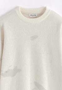 Pull en tricot beige clair avec un col rond, fabriqué en tissu doux. Présente des motifs irréguliers discrets sur le devant. Étiquette de la marque visible.