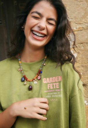 Young woman with dark hair laughs, wearing green "Garden Fete" shirt and colorful beaded necklace, holding hand with ladybug on it.