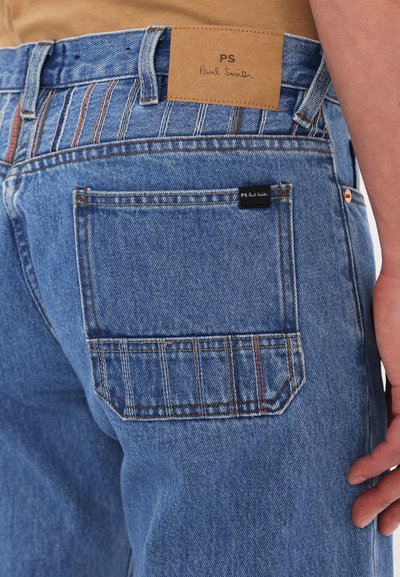 Denim jeans in light blue with a leather tag reading "PS Paul Smith." Features visible stitching and a back pocket with a small logo tag.