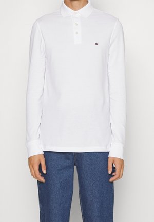 Person iført en hvid langærmet poloshirt med lille logo på brystet og blå jeans, stående mod en ensfarvet baggrund.