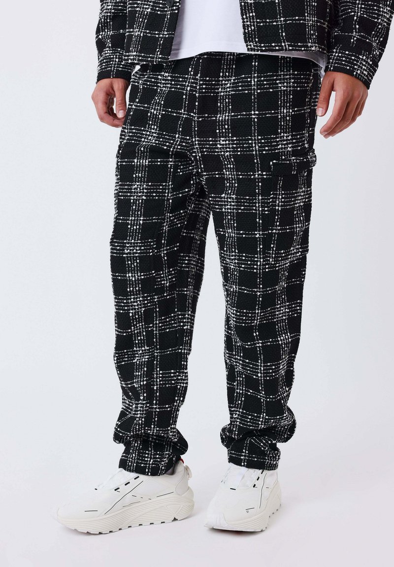 Uomo che indossa pantaloni a scacchi bianchi e neri, giacca coordinata e scarpe da ginnastica bianche spesse, in piedi contro uno sfondo chiaro e semplice.