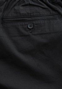Zwarte stof met een achterzak voorzien van een enkele zwarte knop en zichtbare stiksels, die een gladde en matte textuur laat zien.