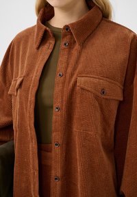 Veste chemise en velours côtelé marron avec un devant à boutons, une poche poitrine unique et un motif en relief texturé. Portée par-dessus un haut vert foncé.