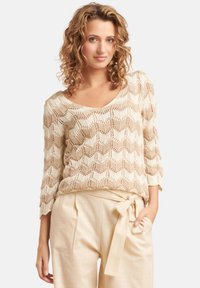 Beige gebreide trui met een chevronpatroon, V-hals en driekwart mouwen, gecombineerd met hooggekraagde, lichtbeige broeken met een strikceintuur.