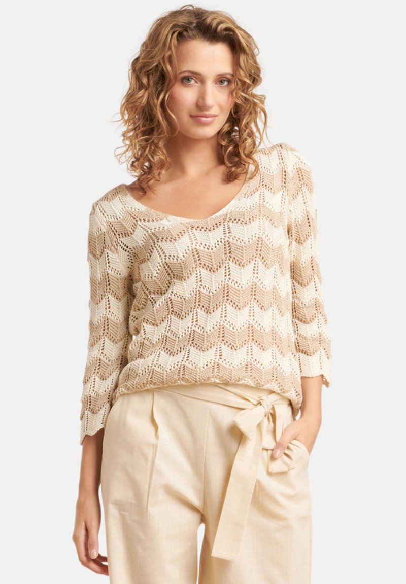 Beige gebreide trui met een chevronpatroon, V-hals en driekwart mouwen, gecombineerd met hooggekraagde, lichtbeige broeken met een strikceintuur.