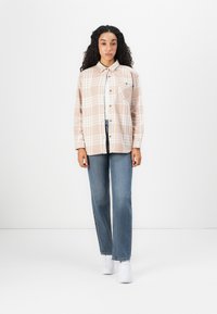 Donna con lunghi capelli ricci, che indossa una camicia a quadri beige, jeans dritti blu e sneakers bianche, in piedi di fronte a uno sfondo bianco.
