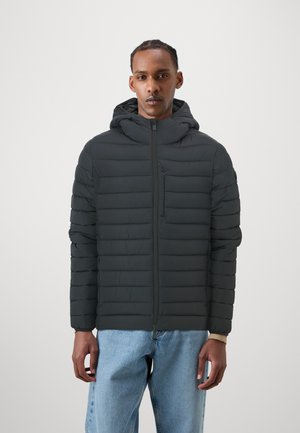 Giacca a vento nera con pattern a cuciture orizzontali, cappuccio attaccato, zip frontale completa e tasca sul petto con zip. Indossata con jeans azzurri chiari.