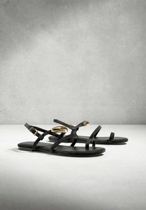 Sandale negre plate cu barete subțiri, catarame la gleznă și accente ovale aurii pe baretele superioare, expuse pe o suprafață simplă, deschisă.