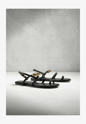 Czarne płaskie sandały z cienkimi paskami, klamrami wokół kostek i złotymi owalnymi akcentami na górnych paskach, prezentowane na jasnej, gładkiej powierzchni.