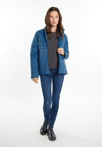 Giacca isolante blu con pannelli testurizzati, chiusura con zip, indossata sopra un maglione grigio, abbinata a jeans scuri e stivaletti neri alla caviglia.