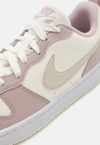 Basket de Nike avec un dessus en cuir rose et crème, des lacets blancs, des accents texturés roses et une semelle en caoutchouc gris clair.