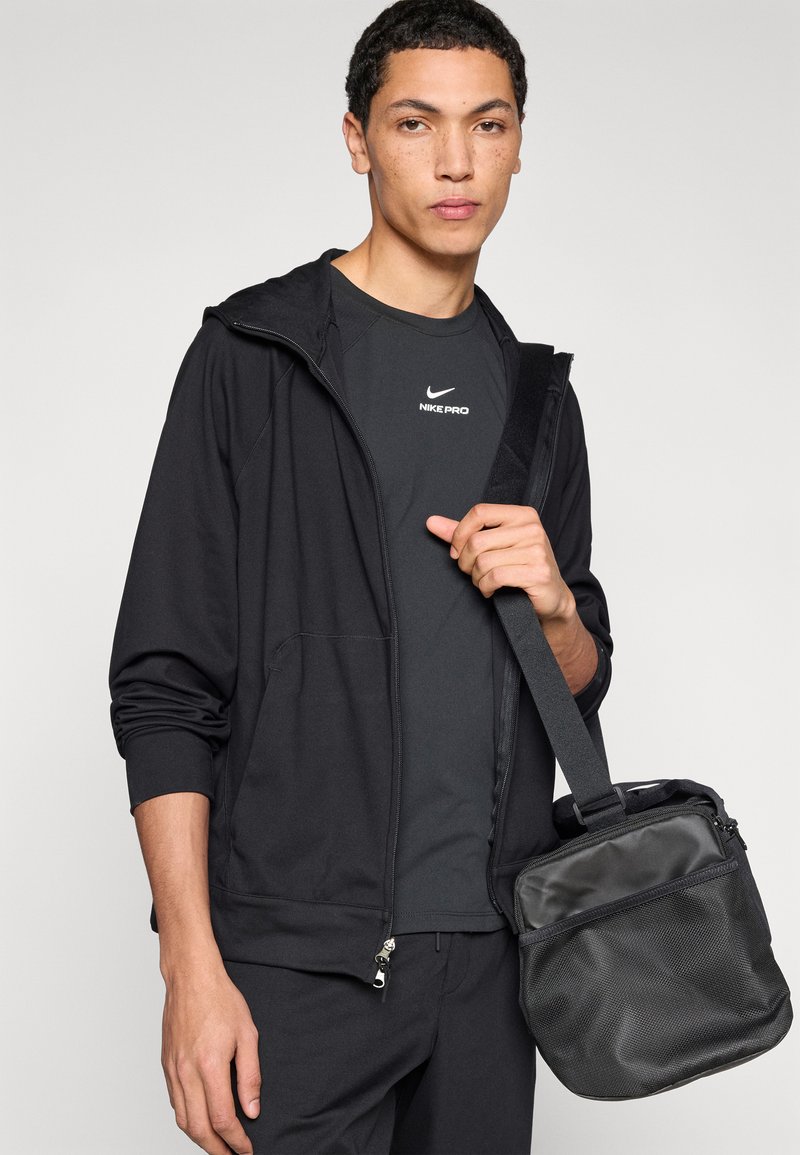 Black hooded jacket made of lightweight fabric, paired with a dark shirt featuring a white logo, holding a black textured bag with a strap.