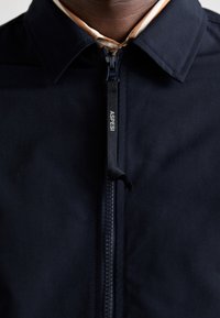 Gros plan d'une veste noire avec un col et une languette de fermeture éclair centrée étiquetée "ASPESI" sur un vêtement intérieur beige.
