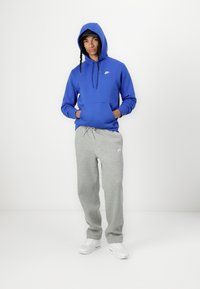 Hoodie azul com um bolso canguru e capuz com cordão, combinado com calças de treino cinzentas claras. Ambos apresentam detalhes de logotipo discretos.