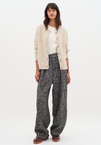 Cardigan en laine beige avec col rond et boutons en bois, associé à un pantalon ample à motif pied-de-poule noir et blanc et des chaussures marron.