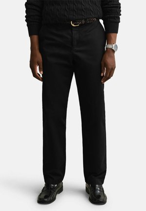 Homme portant un pantalon droit noir, une ceinture noire, un pull noir, des mocassins en cuir noirs et une montre-bracelet argentée sur fond blanc.