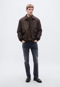 Brun jakke med tekstur og fremre lommer, grønn genser under, blå jeans og svarte sko. Enkel design uten mønstre eller merkbare detaljer.