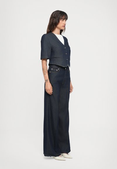 Giacca cropped in denim scuro con maniche corte e chiusura a bottoni, abbinata a jeans wide-leg scuri a vita alta. Scarpe bianche completano il look.