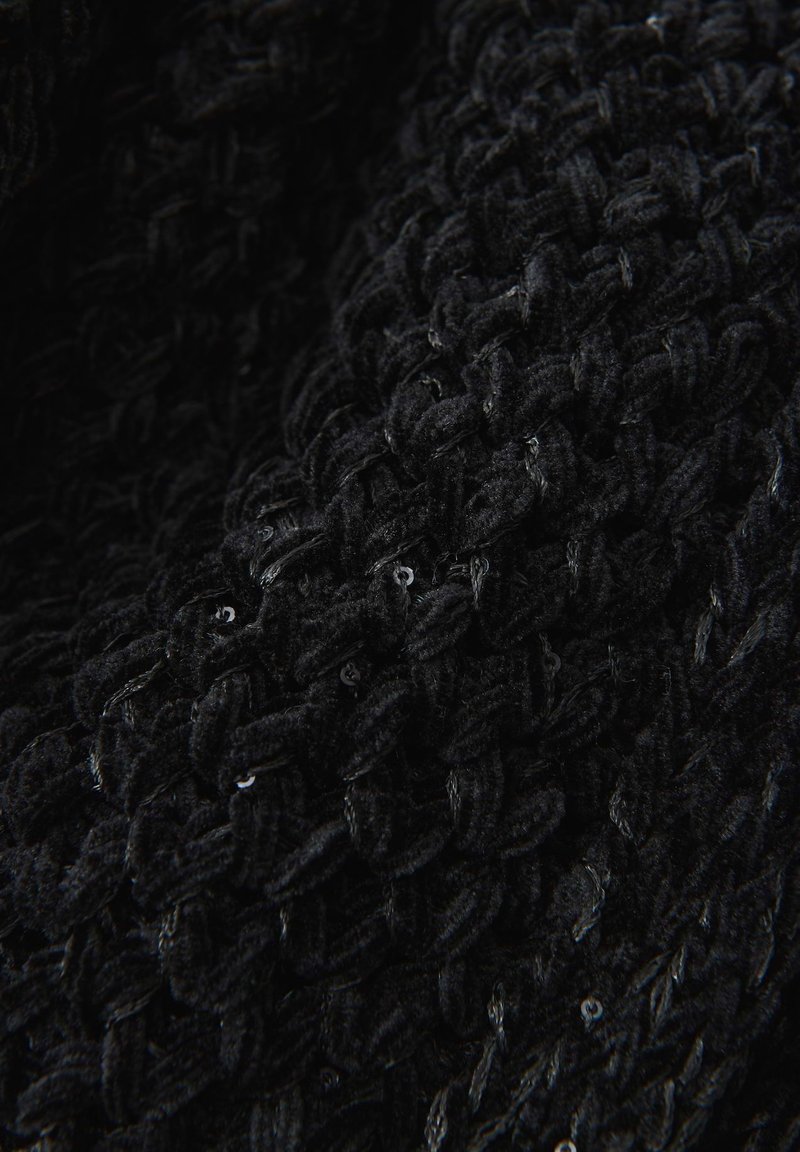 Nahaufnahme von schwarzem, grob gestricktem Stoff mit subtilen, glänzenden Pailletten, die in die strukturierten Schlaufen eingewebt sind.
