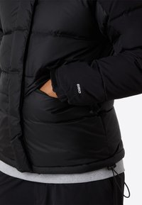 Chaqueta acolchada negra con la mano en el bolsillo con cremallera, manga mostrando bordado "550", llevada sobre un sudadero gris y pantalones negros.