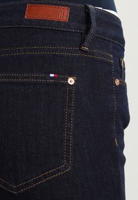Jeans de mezclilla azul oscuro con un parche de cuero en la cinturilla trasera, remaches de cobre y una pequeña etiqueta tricolor en el bolsillo trasero.