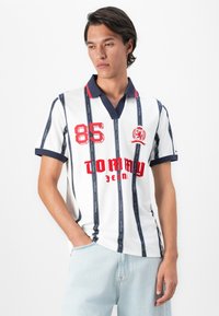 Jonge man in een witte polo met marineblauwe verticale strepen, rode "85" en "Tommy Jeans" tekst, gecombineerd met lichtblauwe jeans, staand tegen een witte achtergrond.