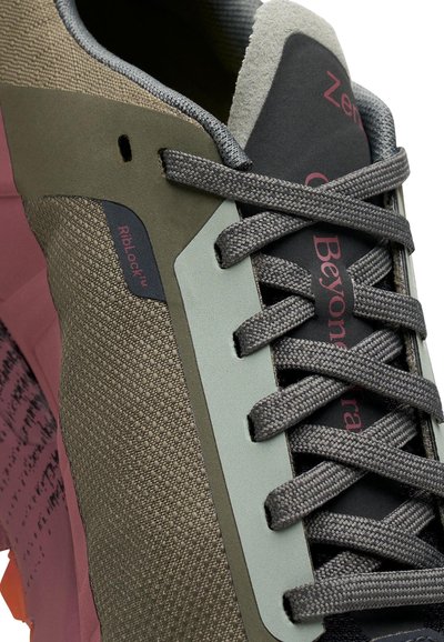 Gros plan sur une sneaker avec des lacets gris, un tissu olive texturé et une semelle rose, affichant "RibLock" et un texte partiellement visible sur la languette.