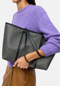 Szara torba tote w geometryczny wzór z czarnymi akcentami, z długimi ramiączkami. Trzymana w zestawieniu z fioletowym swetrem i beżową spódnicą.