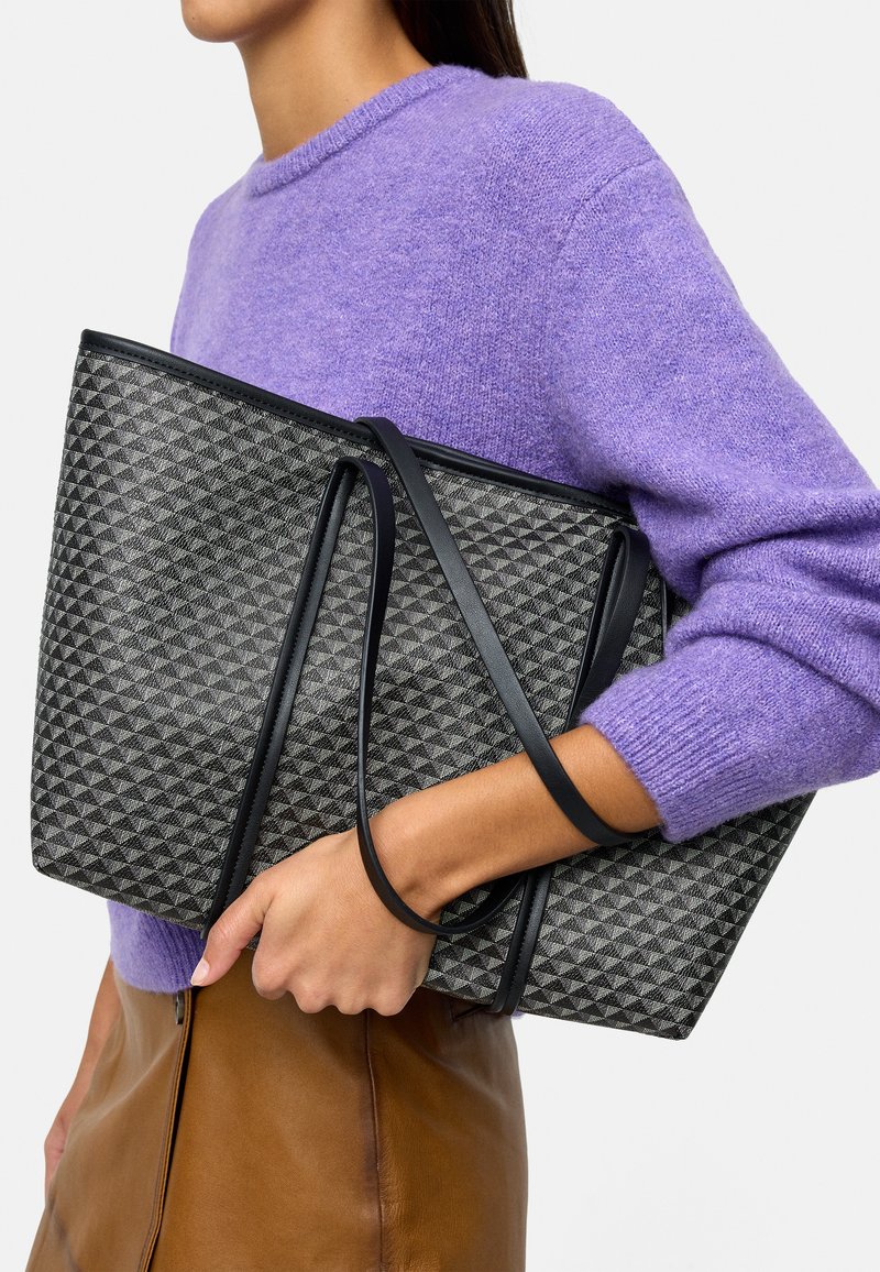 Szara torba tote w geometryczny wzór z czarnymi akcentami, z długimi ramiączkami. Trzymana w zestawieniu z fioletowym swetrem i beżową spódnicą.