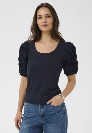 Woman with long brown hair wearing navy puff-sleeve top and blue jeans, standing with one hand in pocket and silver bracelet on wrist.