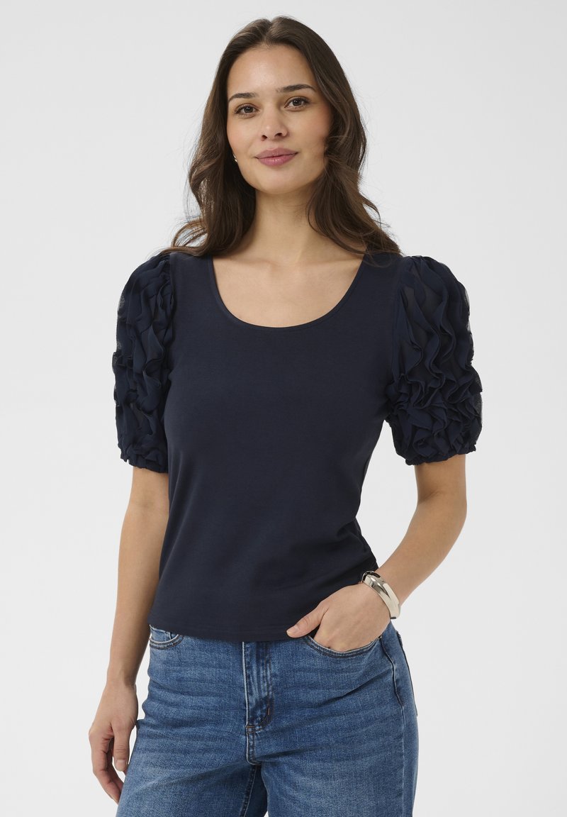 Femme aux longs cheveux bruns portant un haut bleu marine à manches bouffantes et un jean bleu, debout avec une main dans la poche et un bracelet argenté au poignet.