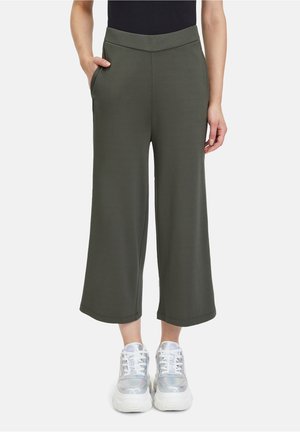 Personne portant un pantalon large court olive, un haut noir et des baskets blanches épaisses, debout avec une main dans la poche.