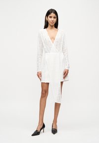 Λευκό δαντελένιο φόρεμα με βαθύ V-λαιμόκοψη και μακριά μανίκια, με ασύμμετρο τελείωμα και ρέον μπροστινό μέρος. Συνδυασμένο με μαύρες γόβες.