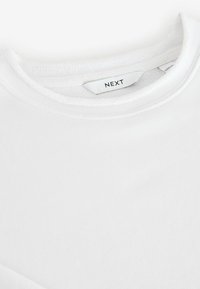 Gros plan d'un t-shirt blanc à col rond montrant l'encolure et une étiquette de vêtement "NEXT" à l'intérieur du col.